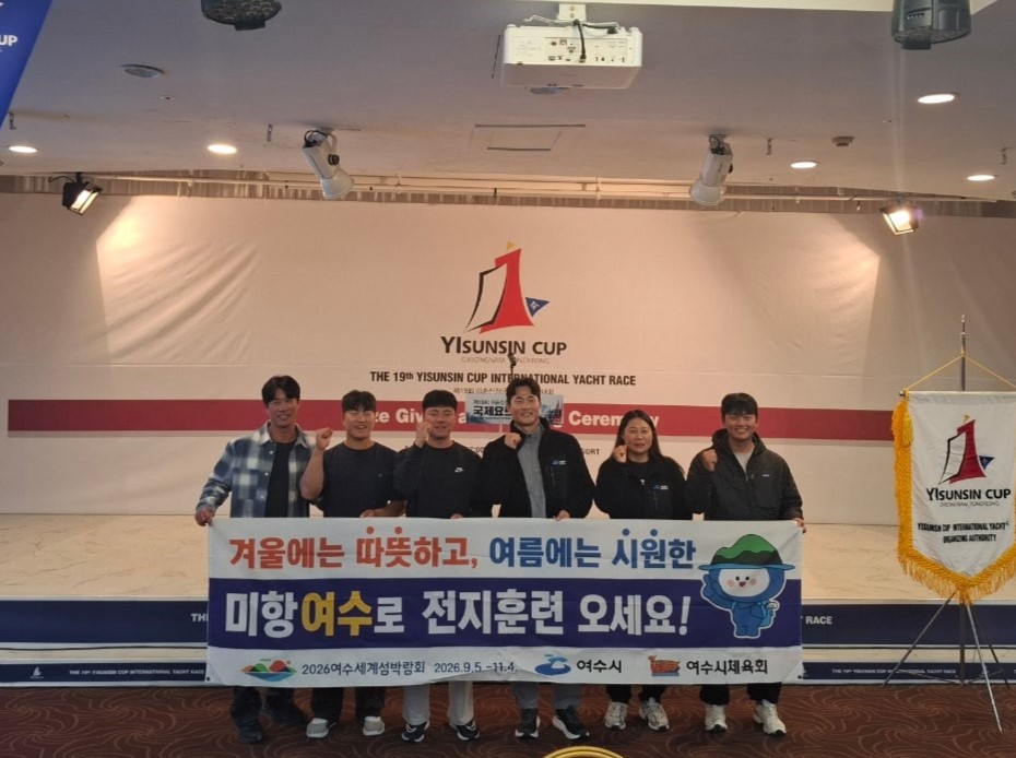 여수시청 요트팀(왼쪽부터 김창주·강형중·이석현·김종승·이경진·성시유 선수)구성원들이 함께 서서 응원 문구가 적힌 현수막을 들고 기념촬영을 하고 있다. 현수막에는 여수에서 전지훈련을 홍보하는 문구가 적혀 있으며 뒤편 무대에는 ‘이순신컵 국제요트대회’ 로고가 보인다.