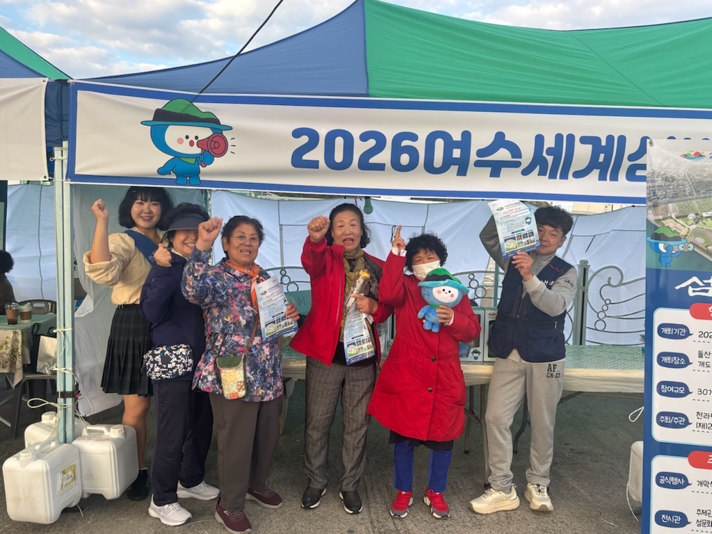 2026여수세계섬박람회 홍보부스에서 주민들이 박람회 안내 전단과 다섬이 인형을 들고 단체 사진을 찍는 모습. 파란색과 초록색 천막 아래 설치된 ‘2026여수세계섬박람회’ 현수막이 보이며, 참가자 대부분이 밝게 웃으며 손을 흔들거나 포즈를 취하고 있다.