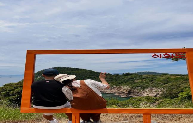 황색 포토프레임 모양의 조형물에 앉아 바다와 숲이 어우러진 섬 풍경을 바라보며 함께 사진을 찍는 두 사람이 어깨를 맞대고 앉아 있고, 뒤로는 푸른 하늘과 자연경관이 펼쳐져 있는 모습.