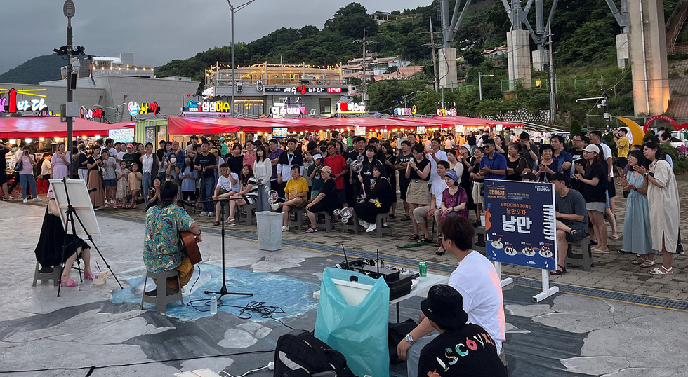 여수 낭만포차 상설 무대에서 밴드가 버스킹을 하고 있고 시민들은  그 무대를 보면서 여가를 보내고 있는 사진이다.