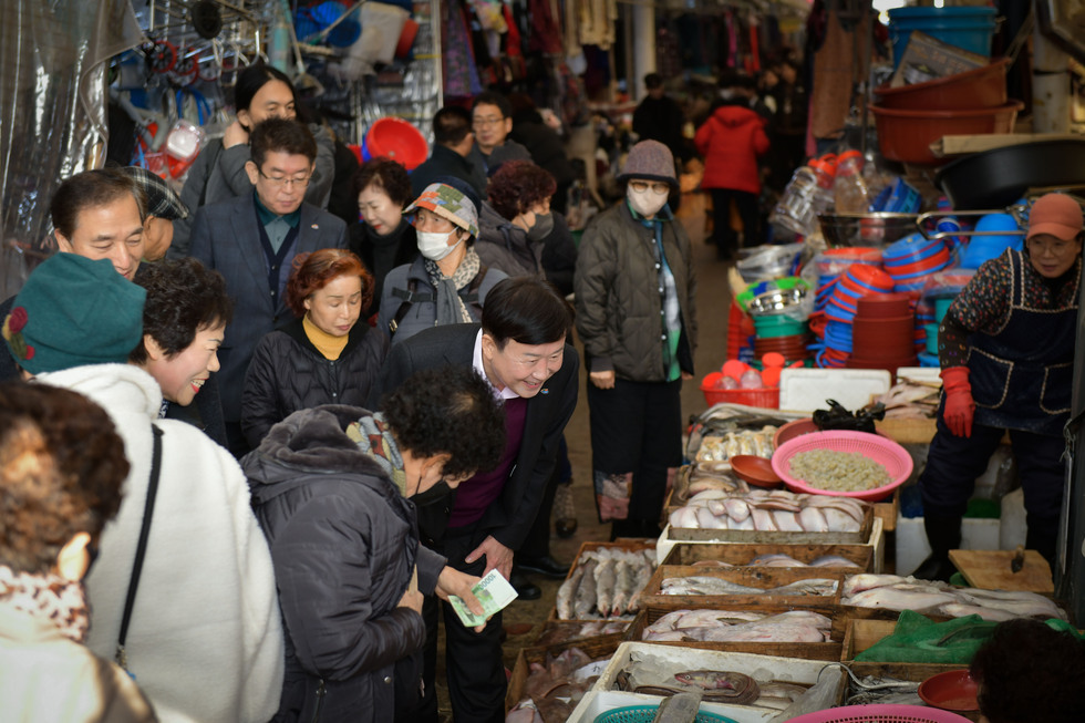 시장 안에서 사람들이 생선과 해산물을 구경하며 대화하고 있는 모습이다. 한 남성이 여성에게 돈을 건네고 있다.