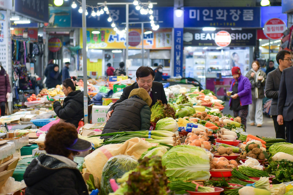 전통 시장에서 여러 종류의 채소와 과일이 진열되어 있으며, 한 남성이 노인 여성과 대화하고 있다.