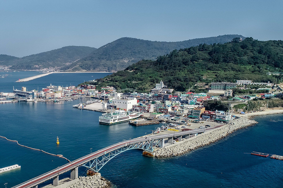 해안가에 위치한 마을과 다리가 보이는 풍경으로, 여러 색상의 건물들이 모여 있는 모습이다. 바다와 산이 배경으로 펼쳐져 있다.