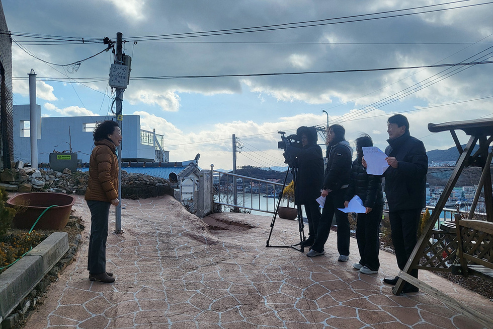 한 여성이 해안가에서 인터뷰를 받고 있으며, 카메라를 든 촬영팀이 그녀를 촬영하고 있다. 배경에는 바다와 구름이 보인다.