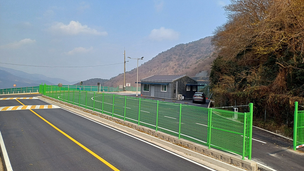 도로 옆에 위치한 작은 건물과 초록색 펜스가 있는 장면으로, 배경에는 산이 보인다.