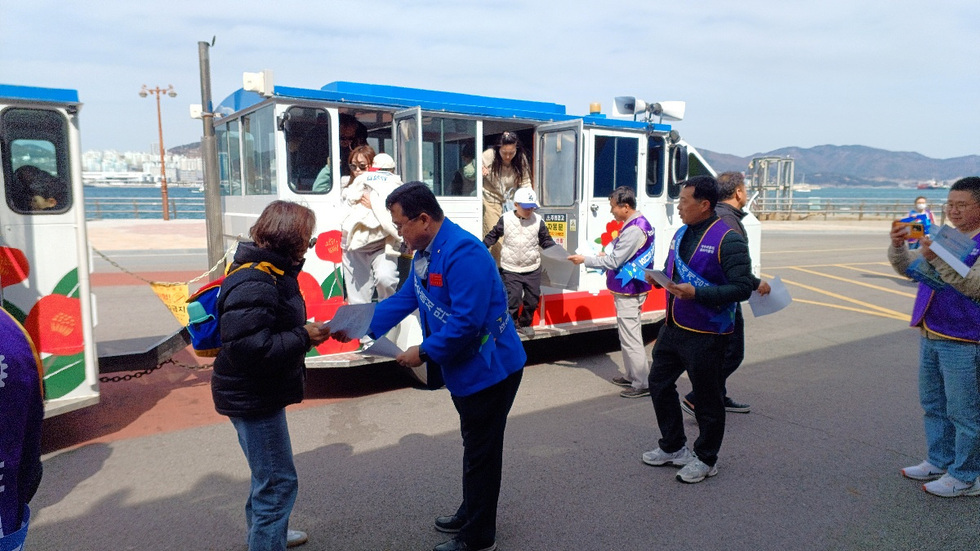 사람들이 해안가에서 차량에서 내리며 안내문을 받고 있는 장면이다. 여러 사람이 차량 주위에서 활동하고 있다.