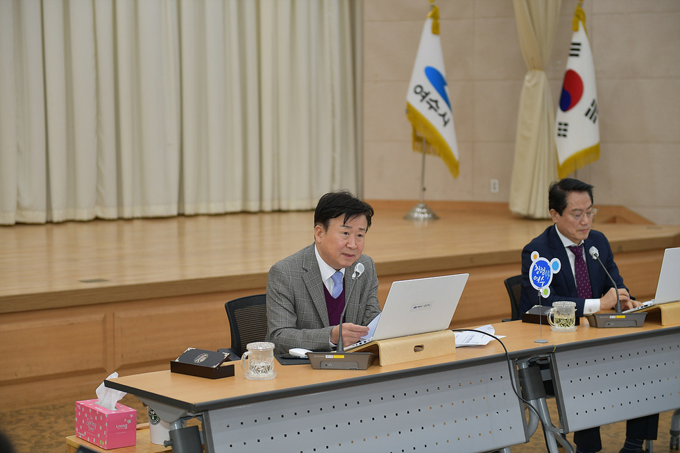 여수시청에서 회의를 진행하는 모습으로, 한 남성이 노트북을 보며 발언하고 있고, 다른 남성은 서류를 보고 있다. 배경에는 여수시와 대한민국 국기가 보인다.