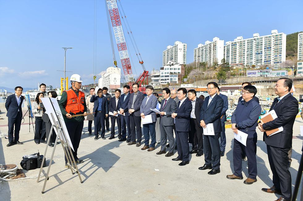 한 남성이 안전모를 착용하고 구명조끼를 입고 있으며, 여러 명의 사람들이 그를 바라보며 서 있다. 배경에는 크레인과 아파트 건물이 보인다.