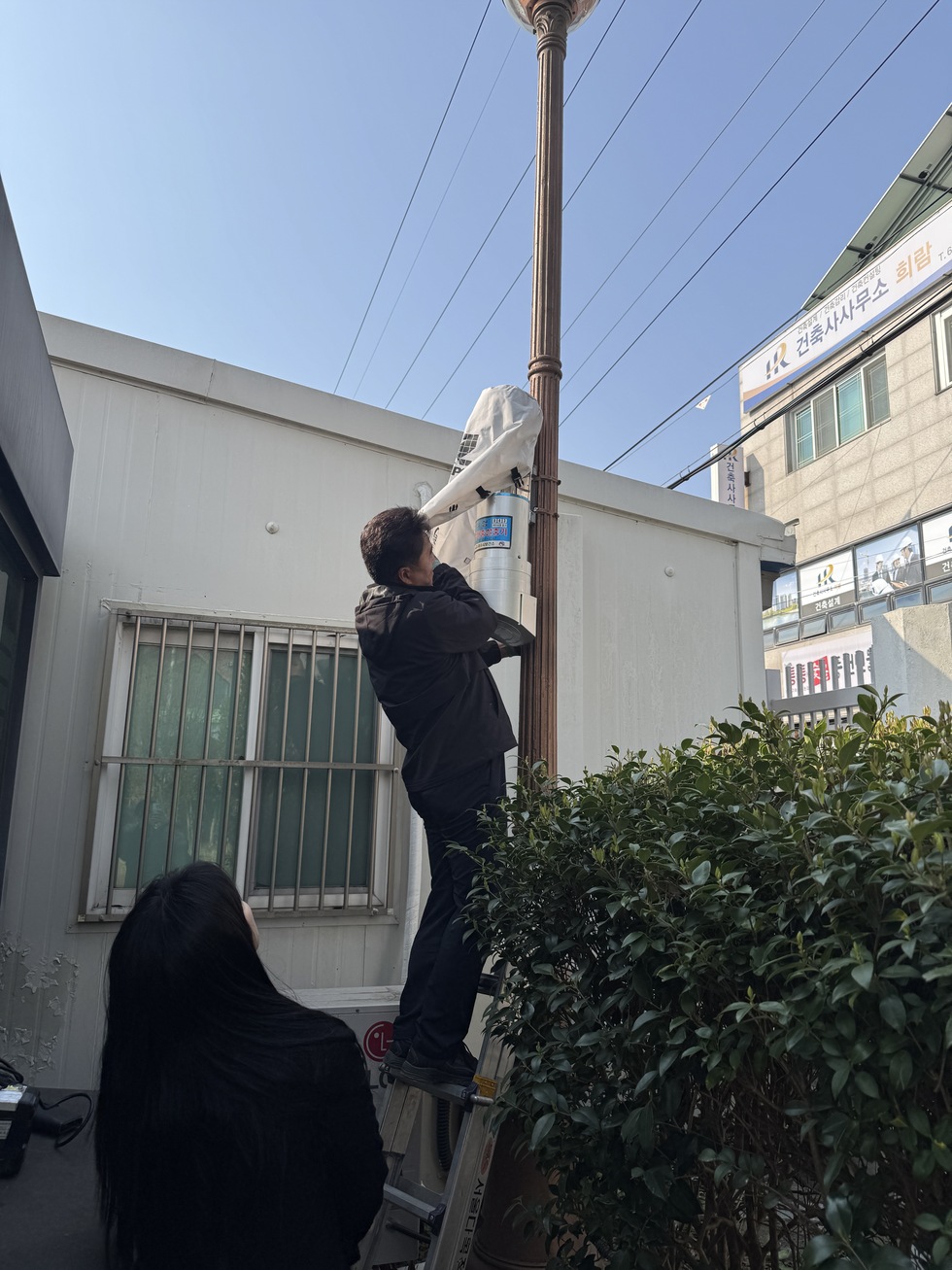 여수시 관계자가 사다리를 타고 가로등 기둥에 설치된 태양광 패널과 장치를 점검하는 모습. 아래에서는 또 다른 직원이 안전을 위해 지켜보고 있다.