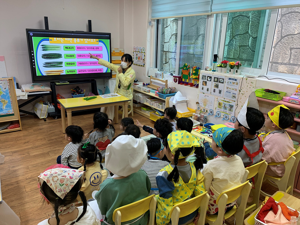 교사가 아이들에게 다양한 오이의 종류를 설명하고 있는 유치원 교실의 모습이다. 아이들은 교사의 설명을 듣고 있다.