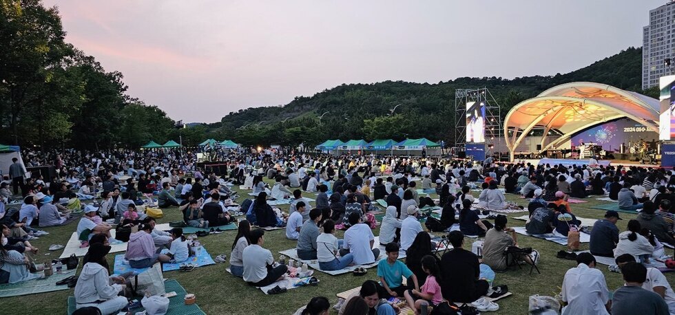 많은 사람들이 야외 공연장에서 음악을 감상하고 있는 모습으로, 무대에는 공연자가 서 있고 배경에는 공연 관련 정보가 적힌 현수막이 보인다.