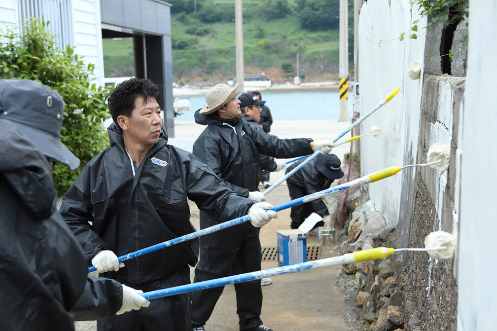 여러 사람이 검은 작업복을 입고 벽을 페인트칠하고 있는 모습이다. 배경에는 바다와 산이 보인다.