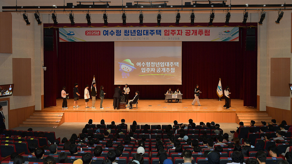 여수문화홀에서 2025년 여수형 청년임대주택 입주자 공개추첨이 진행되고 있으며, 무대에는 여러 사람이 서 있고, 심사위원들이 앉아 있다.