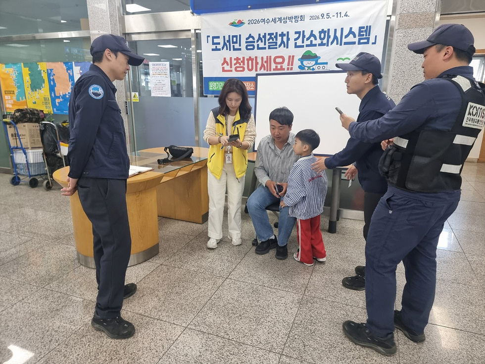 도서민 승선절차 간소화 시스템에 대해 안내하는 장면으로, 직원이 가족과 대화하고 있으며, 배경에는 관련 현수막이 걸려 있다.