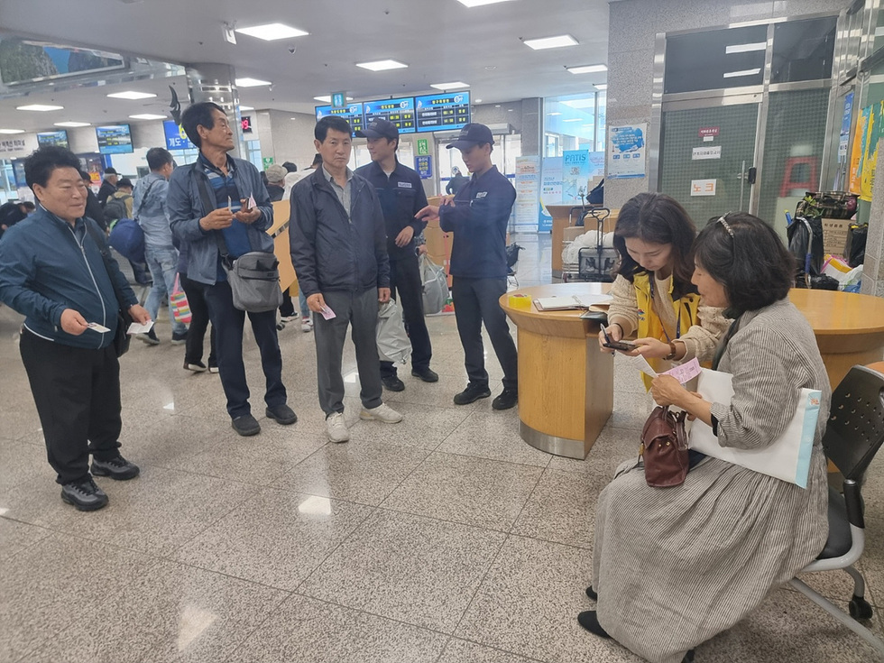 사람들이 대기 중인 장소에서 여러 명이 모여 이야기를 나누고 있다. 두 여성은 스마트폰을 보며 대화하고 있으며, 주변에는 다른 사람들이 대기하고 있다.