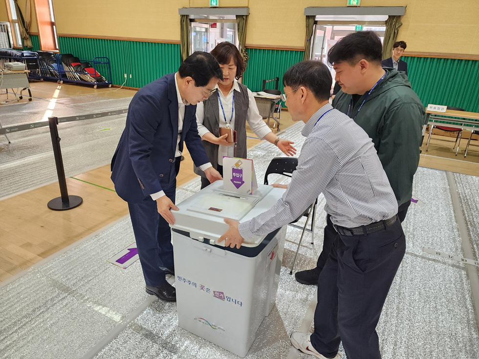 투표소에서 여러 사람이 투표함을 살펴보며 대화하는 장면이다.