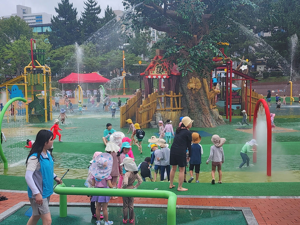 어린이들이 물놀이를 즐기는 공원에서 여러 명의 아이들이 분수와 놀이시설 주변에서 놀고 있다. 한쪽에서는 성인이 아이들을 지켜보고 있다.