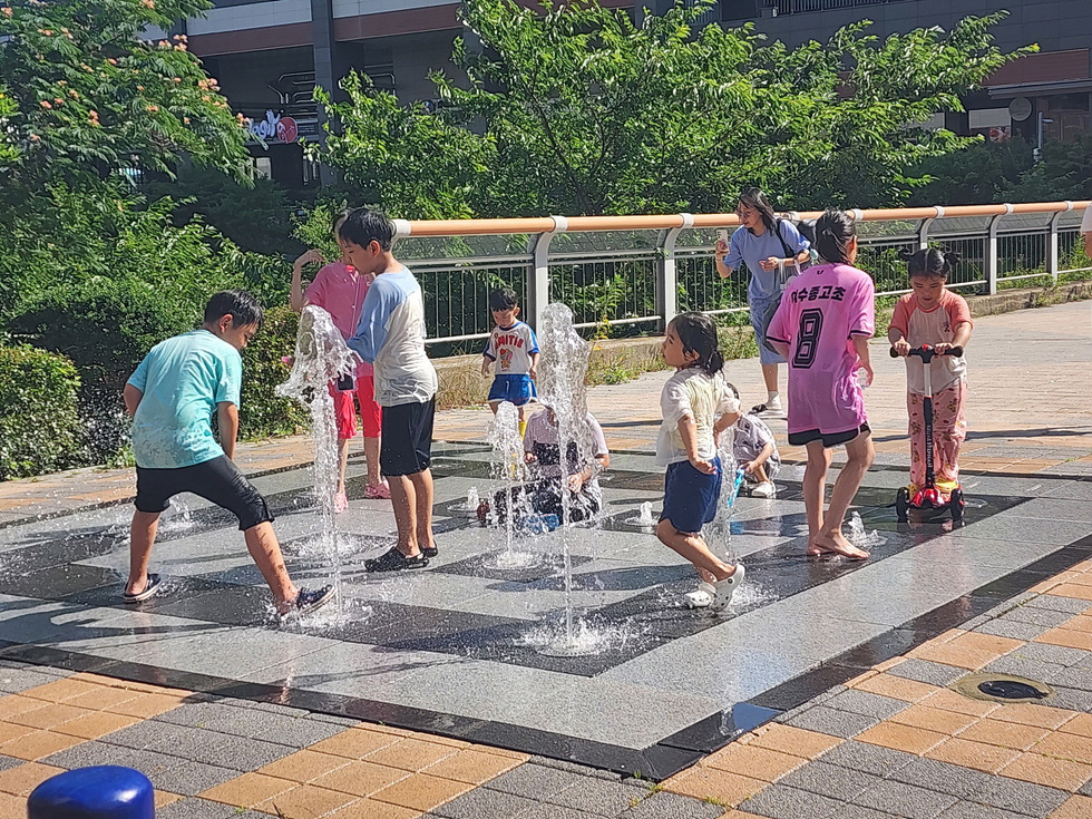 여름날 아이들이 분수대에서 물놀이를 하며 즐거운 시간을 보내고 있다. 몇몇 아이들은 물줄기 아래에서 뛰어놀고 있고, 다른 아이들은 킥보드를 타고 있다.