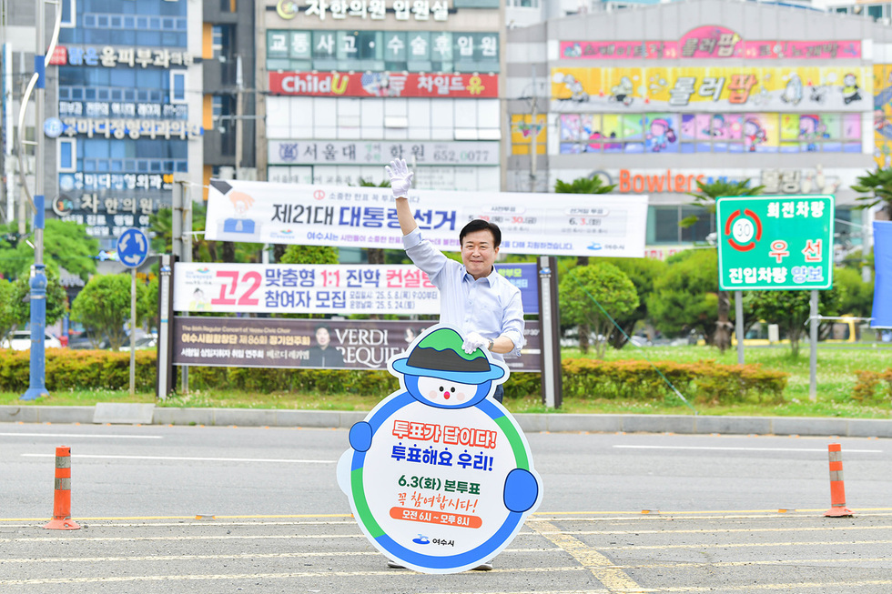 한 남성이 투표를 독려하는 캐릭터 모양의 간판을 들고 서 있다. 배경에는 제21대 대통령선거와 관련된 현수막이 보인다.