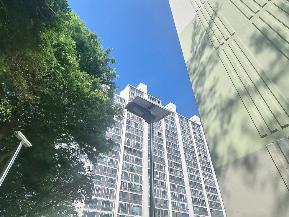 푸른 하늘 아래 고층 아파트와 가로등이 보이는 장면으로, 주변에 나무가 우거져 있다.