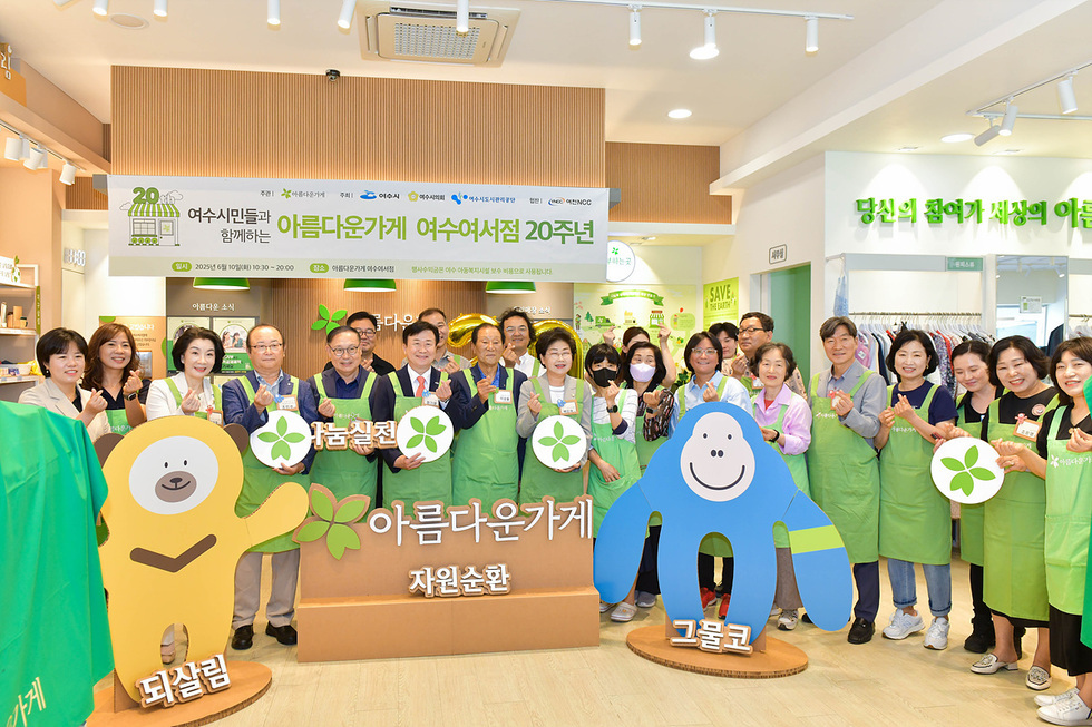 여수여점 20주년 기념 행사에서 다양한 사람들이 모여 기념사진을 찍고 있다. 참석자들은 초록색 앞치마를 착용하고 있으며, 배경에는 행사 관련 현수막과 캐릭터가 보인다.