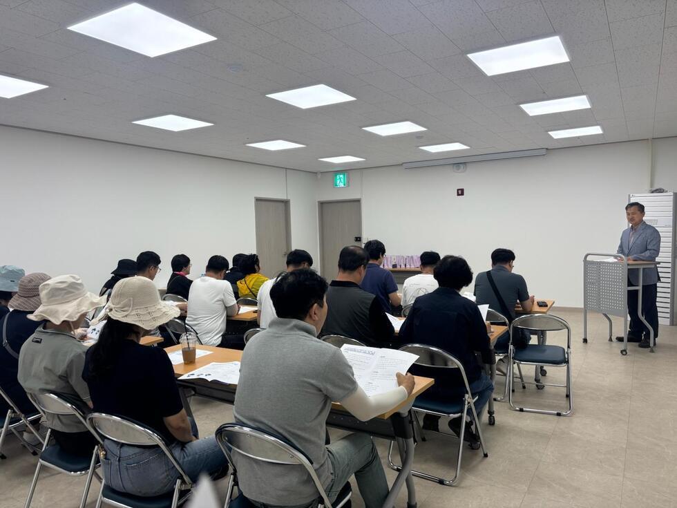 강사가 강의 중인 모습으로, 청중은 테이블에 앉아 강의를 듣고 있다. 방 안에는 여러 명의 사람들이 있으며, 일부는 모자를 쓰고 있다.