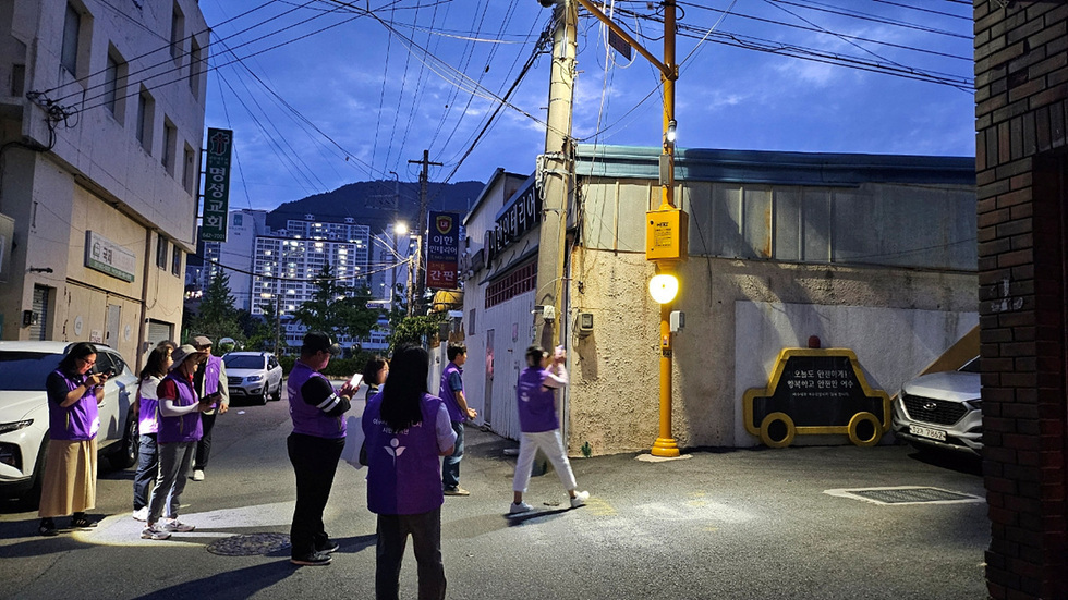 밤에 사람들이 보라색 조끼를 입고 거리에서 활동하고 있으며, 안전과 건강을 강조하는 메시지가 적힌 표지판이 보인다.