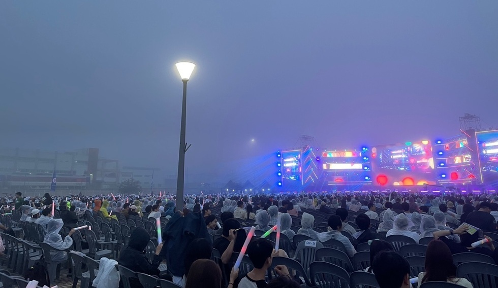 안개가 낀 야외 공연장에서 많은 관객들이 비옷을 입고 있으며, 손에 형광봉을 들고 있다. 무대에는 화려한 조명이 설치되어 있다.