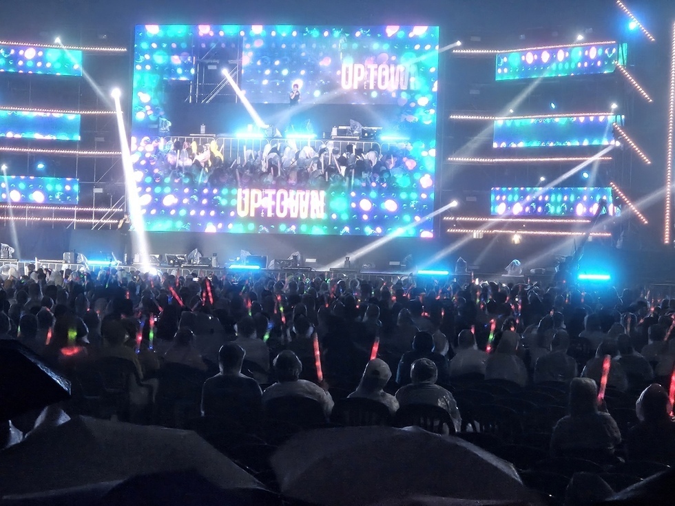UPTOWN이라는 이름의 공연이 진행되고 있는 대형 스크린과 관객들이 보이는 장면이다. 관객들은 다양한 색상의 응원봉을 들고 있다.