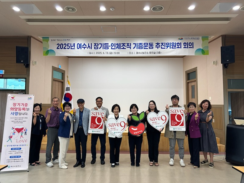 여수시청소년 회의실에서 2025년 장기기증 추진위원회 회의가 열리고 있으며, 참석자들이 장기기증을 홍보하는 포스터와 배너를 들고 있다.