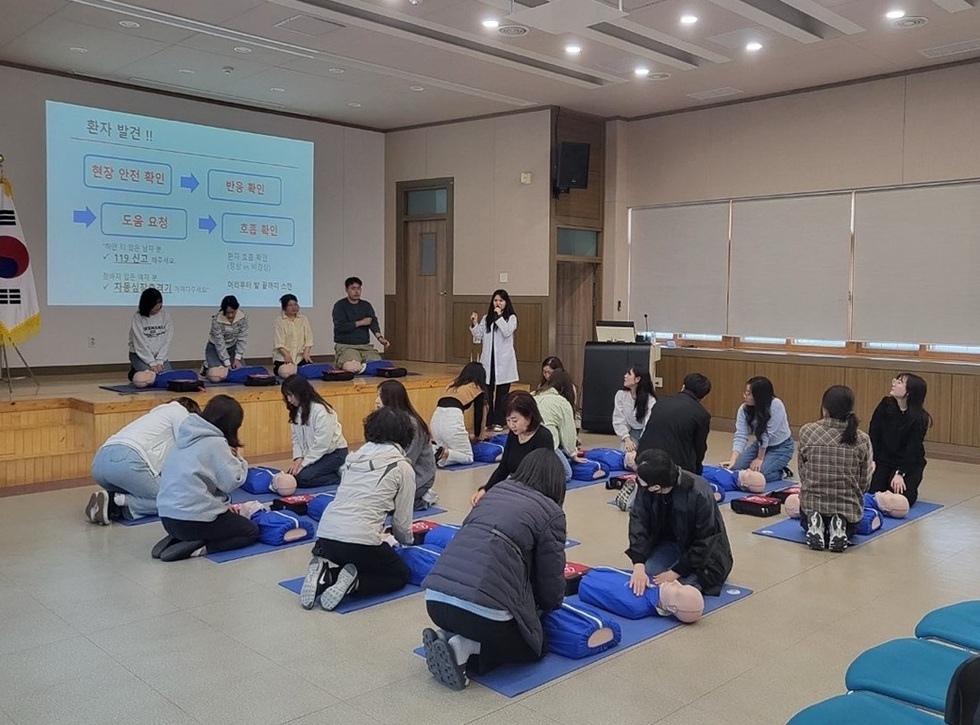여러 사람이 심폐소생술 교육을 받고 있는 장면으로, 강사가 설명하고 있으며 참가자들은 인형을 사용해 실습하고 있다.