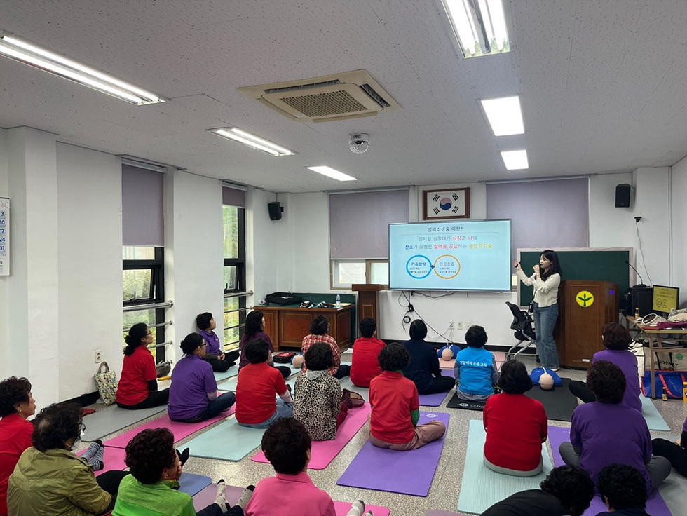 여성이 강의 중이며, 청중은 바닥에 앉아 있다. 화면에는 신체 노화와 건강에 대한 내용이 표시되어 있다.