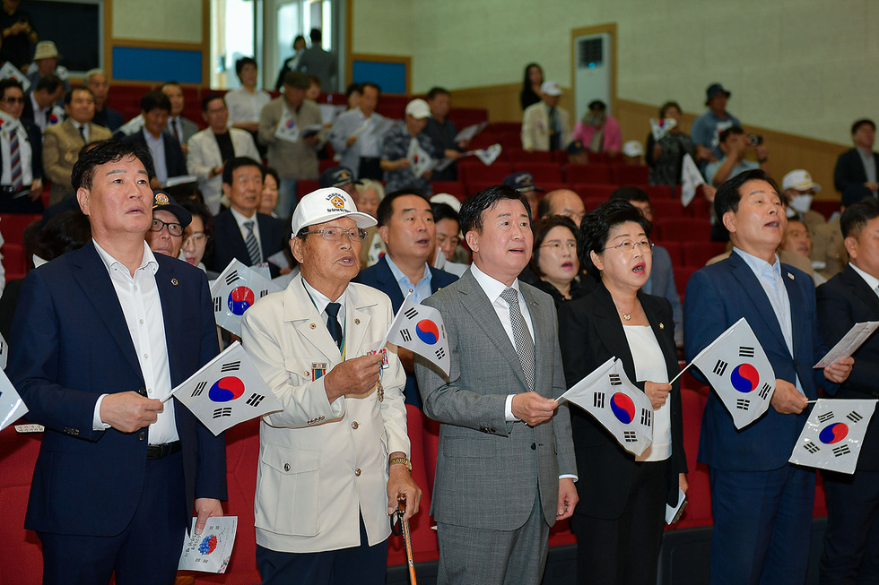 여러 사람들이 태극기를 들고 노래를 부르며 기념행사에 참여하고 있는 모습이다. 행사장에는 많은 관중이 모여 있다.