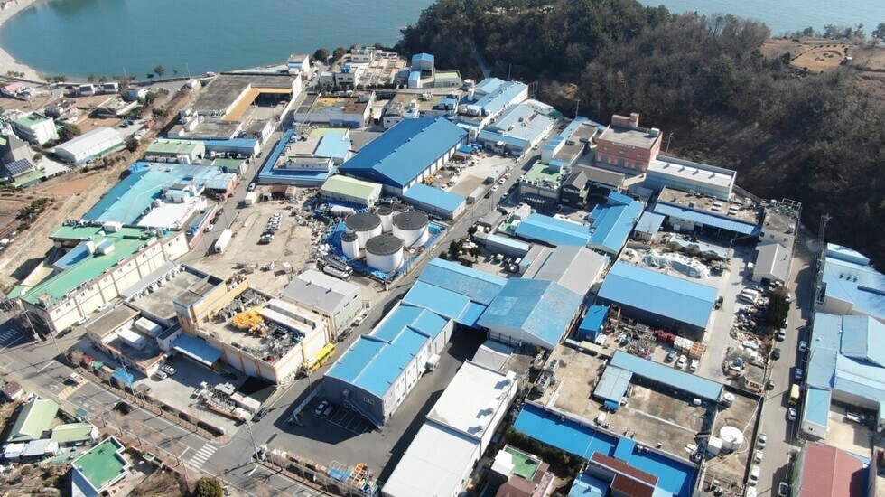 바다 근처의 산업 단지 전경으로 여러 개의 건물과 주차된 차량들이 보인다. 대부분의 건물 지붕은 파란색이며, 주변에 도로와 나무들이 있다.