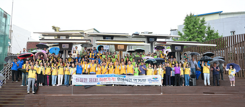 여수세계섬박람회 관련 행사에서 참가자들이 노란 조끼를 입고 단체 사진을 찍고 있다. 현수막에는 친절한 응대와 적정가격, 위생적인 먹거리에 대한 내용이 적혀 있다.