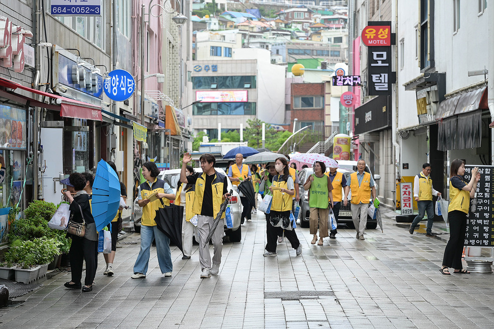 사람들이 노란 조끼를 입고 거리에서 활동하고 있으며, 일부는 우산을 쓰고 있다. 상점 간판과 거리의 모습이 보인다.