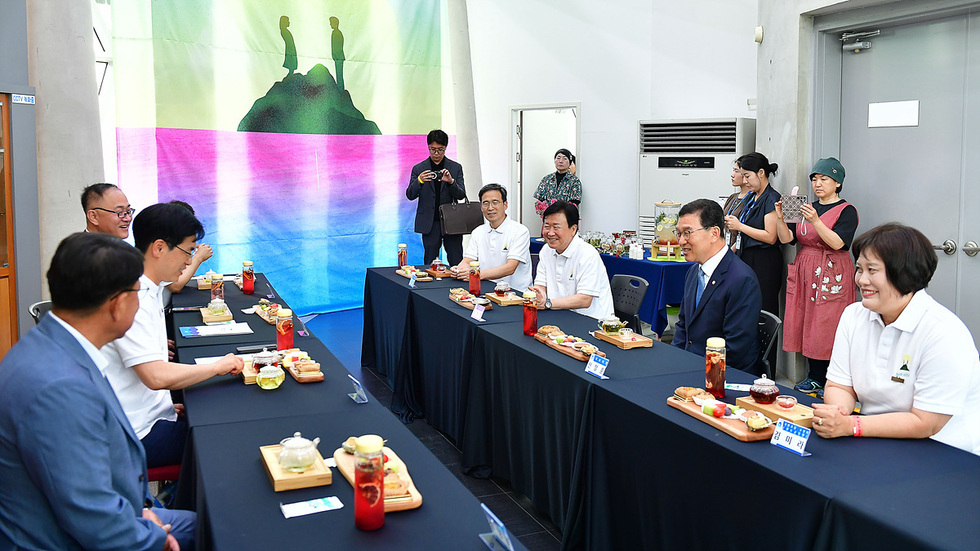 여러 명의 사람들이 테이블에 앉아 음식을 나누며 대화하고 있는 장면이다. 배경에는 색색의 천이 걸려 있다.