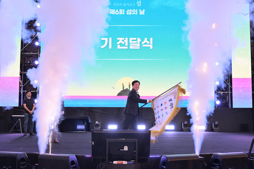 행사에서 한 남성이 기념 깃발을 들고 있으며, 배경에는 제6회 섬의 날을 알리는 대형 화면이 있다.
