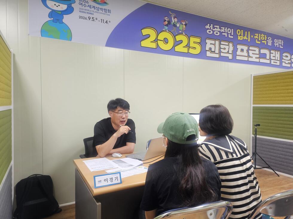 이경기라는 이름의 남성이 두 명의 여성과 대화하고 있으며, 2025 진학 프로그램 운영에 대한 설명을 하고 있다. 배경에는 2026 여수 세계섬박람회 관련 현수막이 걸려 있다.