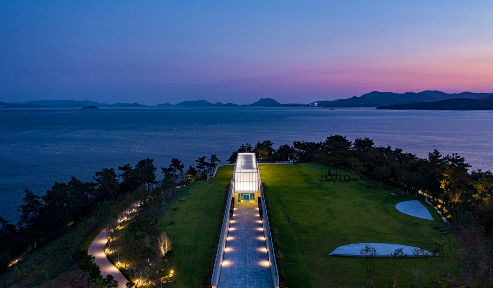 저녁 노을이 지는 바다를 배경으로 한 현대적인 건물이 보이는 풍경이다. 건물 앞에는 넓은 잔디밭과 조명이 설치된 산책로가 있다.