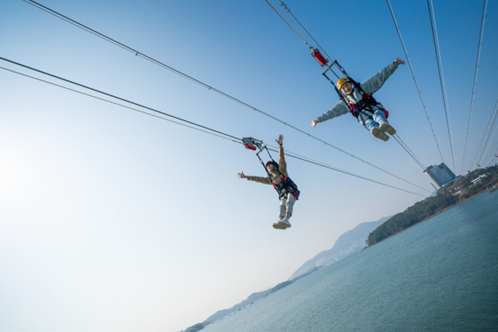 두 사람이 짚라인을 타고 하늘을 나는 모습으로, 배경에는 바다와 산이 보인다.