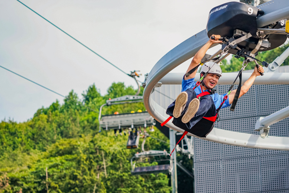 한 남성이 짚라인을 타고 즐거운 표정으로 공중을 가로지르고 있다. 배경에는 나무와 케이블카가 보인다.