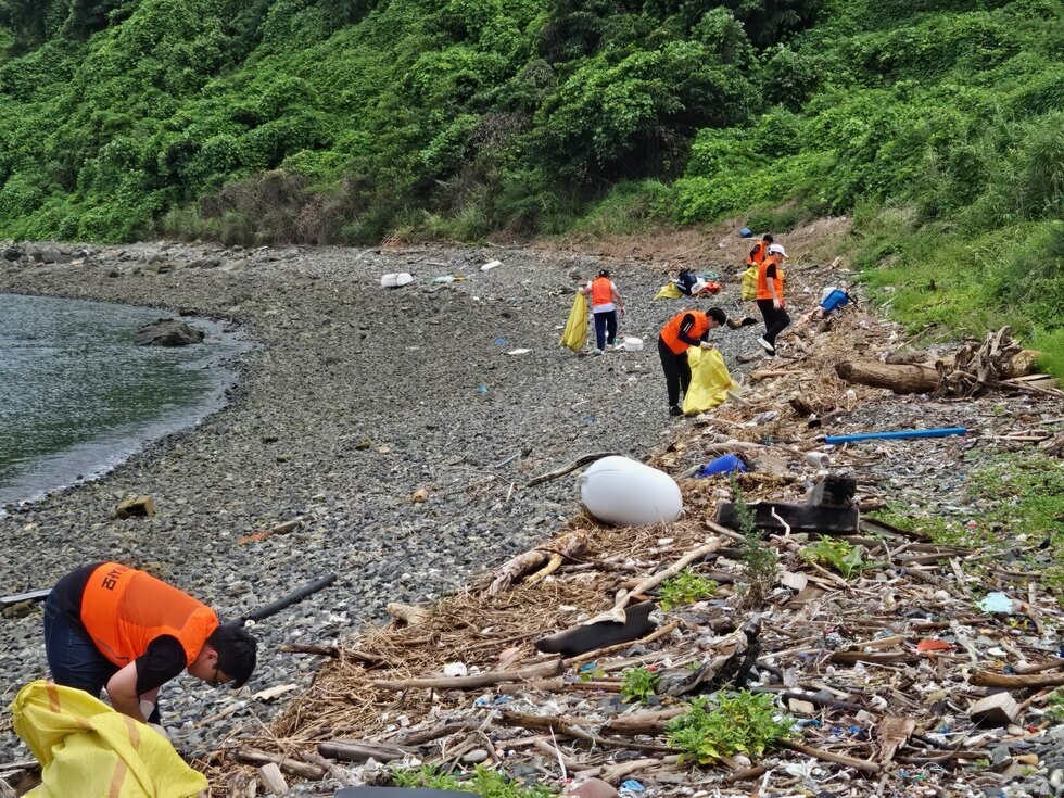 해변에서 자원봉사자들이 쓰레기를 줍고 있는 모습이다. 일부는 노란 봉투를 들고 있으며, 주변에는 많은 쓰레기가 널려 있다.