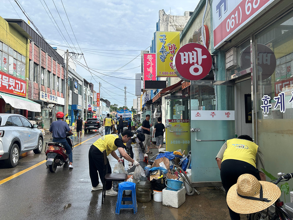 사람들이 도로에서 쓰레기를 치우고 있는 모습으로, 상점들이 줄지어 있는 거리에서 자원봉사자들이 청소 작업을 하고 있다.