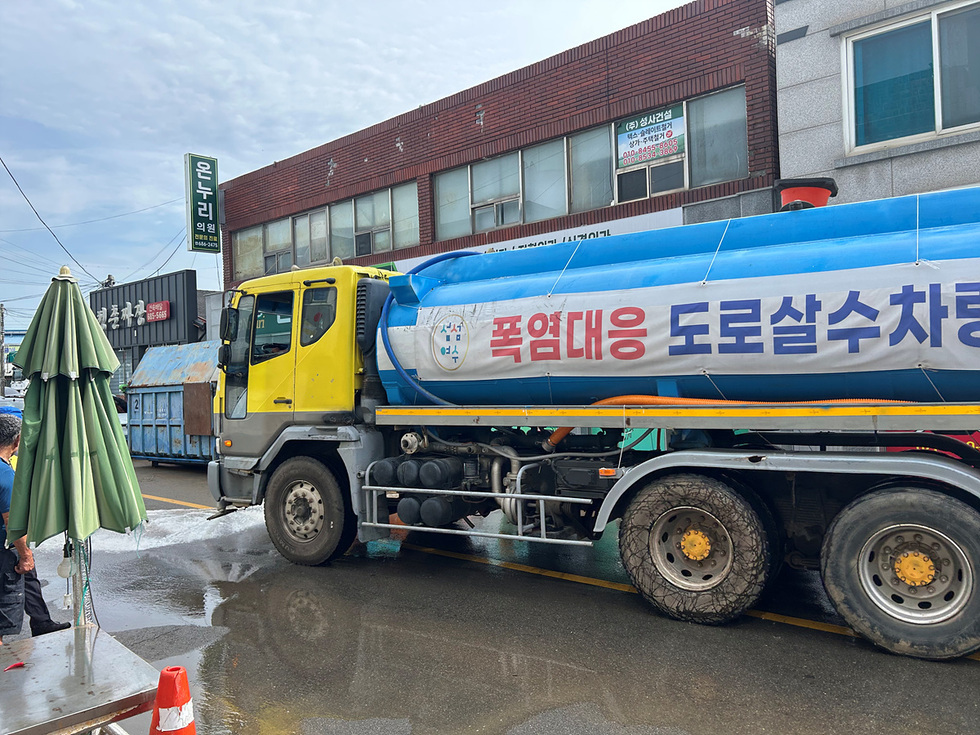 도로에서 폭염 대응을 위해 물을 뿌리고 있는 도로 살수 차량이 보인다. 차량 옆에는 파라솔이 세워져 있다.