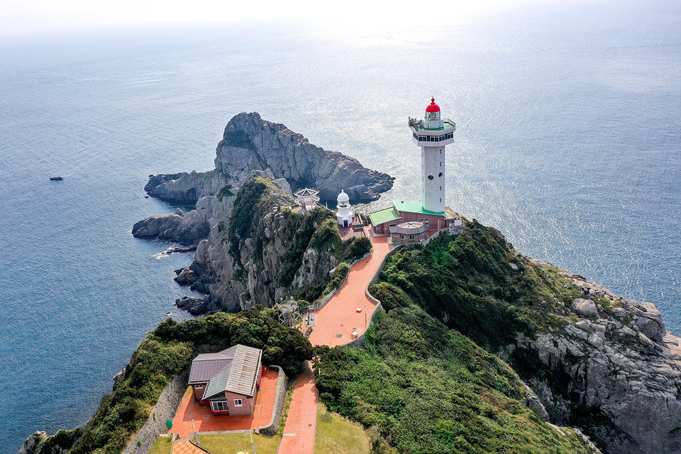 바다 위 절벽 끝자락에 자리한 거문도 등대와 주변 건물, 울창한 녹지와 기암괴석이 어우러진 절경이 펼쳐진 모습으로, 국가 자연유산(명승)으로 지정 예고된 거문도 수월산 일원의 풍경