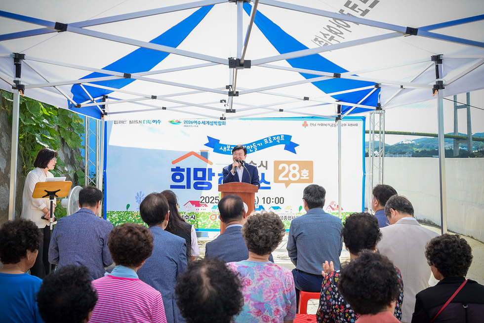 파란 천막 아래에서 진행된 희망하우스 28호 준공 현판식에서 연단에 선 사회자가 마이크를 잡고 인사말을 전하며 앞줄에 모인 지역 주민과 관계자들이 경청하고 있는 모습.