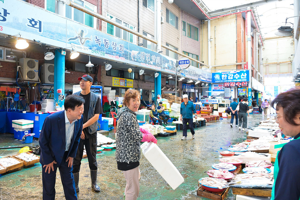 여수시장이 교동시장 골목에서 상인들과 함께 시장 상황을 둘러보며 이야기를 나누는 장면. 바닥에는 수산물이 진열되어 있고, 여러 상인과 시민들이 분주히 움직이고 있다.