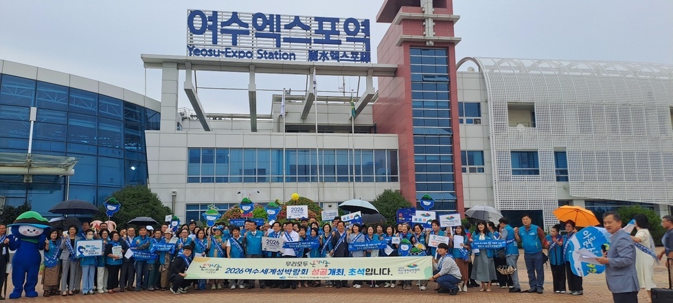 여수엑스포역 앞에서 시민들이 2026여수세계섬박람회 홍보 캠페인에 참여하고 있다. 시민들은 온기나눔 2026년 여수세계섬박람회 성공개최 초석입니다 슬로건이 적힌 현수막을 들고 있다.