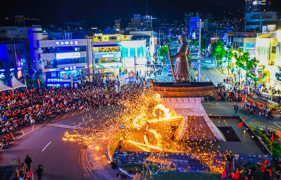 이순신광장 일대에서 펼쳐진 여수밤바다 축제 현장. 이순신 장군 동상 앞에서 불꽃과 불길이 터오르며 관람객들이 도로 주변을 가득 메우고 있다. 도시의 네온사인 불빛과 함께 생동감 넘치는 밤 풍경이 펼쳐진다